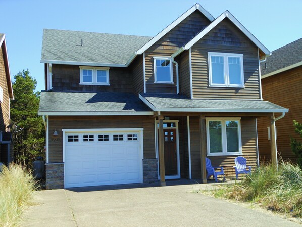 A two-story house with a garage, a front door, and a small porch with two Adirondack chairs.