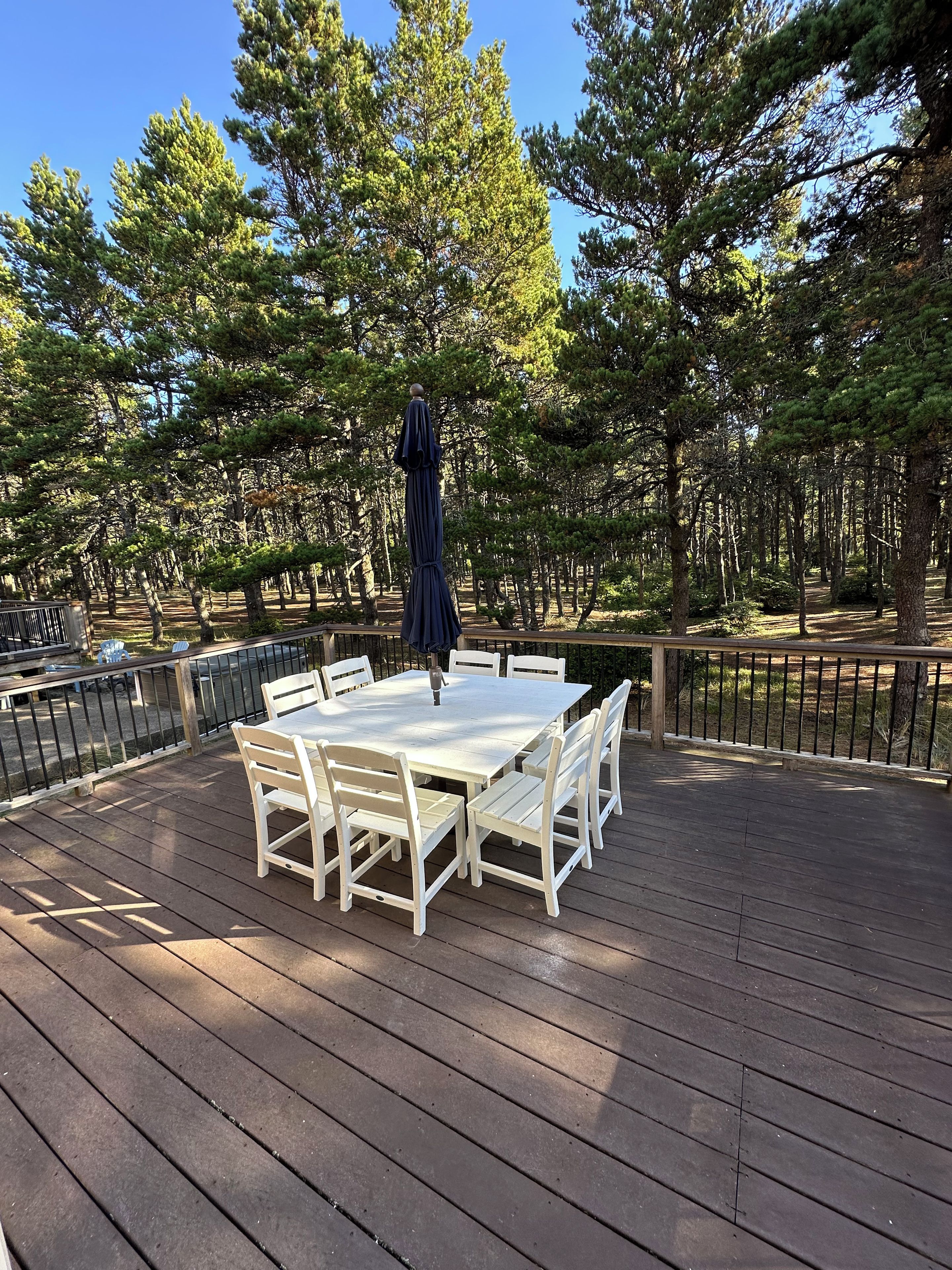 A wooden deck with a white table and chairs, surrounded by a metal railing, set in a forested area.