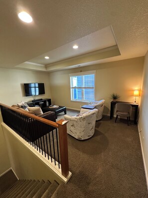 A living room with a flat-screen TV, a fireplace, a window with blinds, a lamp, and a staircase.