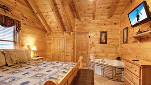 A wooden cabin bedroom with a bed, a nightstand, a lamp, a television, and a stone fireplace. - !BEST LOCATION! RESERVE NOW FOR SPRING BREAK AND SUMMER! BEARFOOT CORNER CABIN!  (Pigeon Forge)