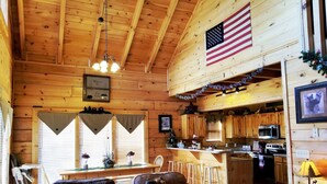 A cozy cabin interior with a wooden ceiling, a dining area with a table and chairs, a kitchen with wooden cabinets and appliances, and a living room with a leather sofa and a fireplace. - !BEST LOCATION! RESERVE NOW FOR SPRING BREAK AND SUMMER! BEARFOOT CORNER CABIN!  (Pigeon Forge)