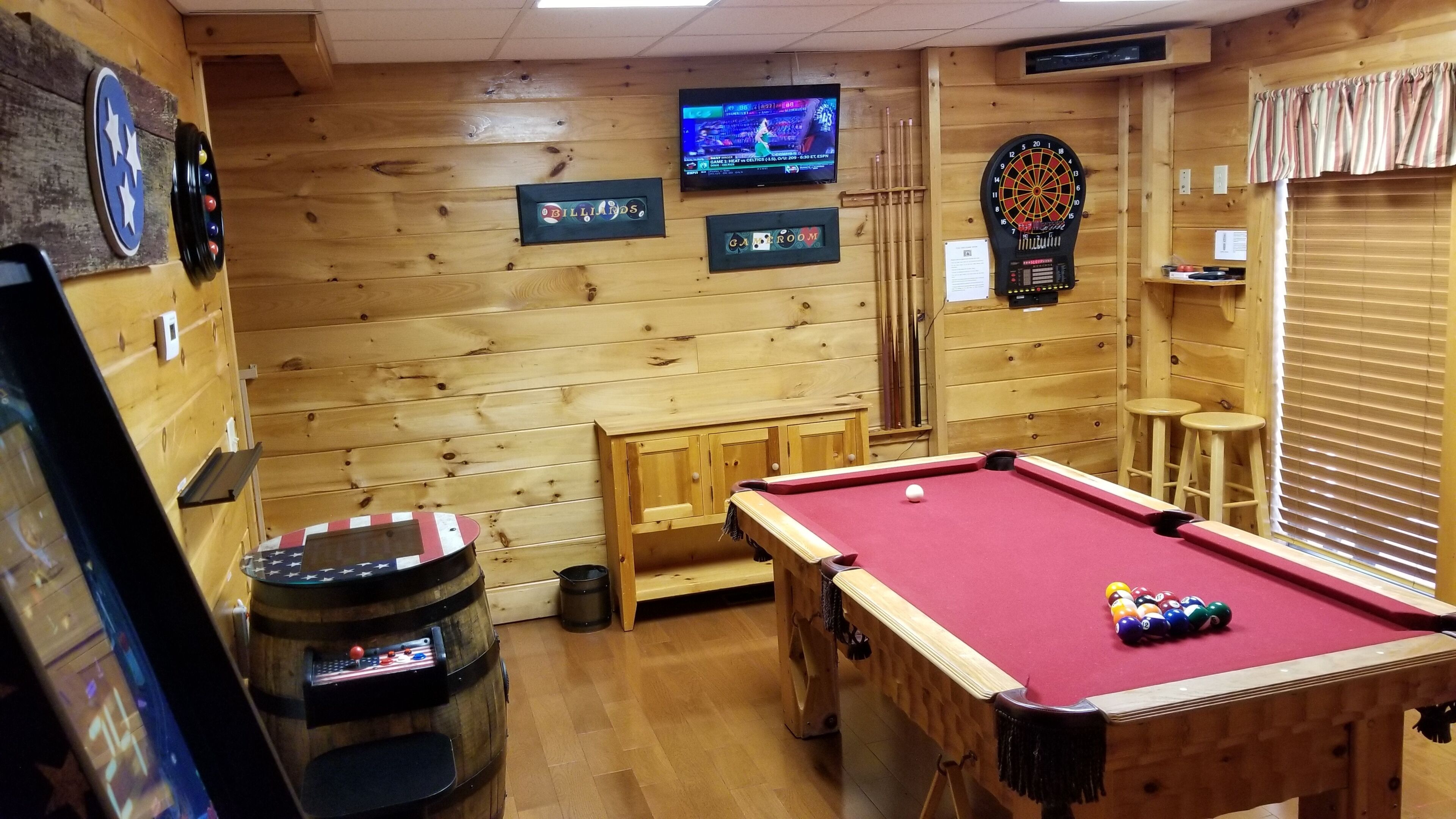 A billiards room with a pool table, a dartboard, and a television displaying a sports event.