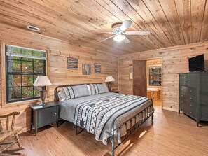 A wooden cabin bedroom with a bed, two bedside lamps, a ceiling fan, and a dresser. - Country Charm D - Cozy Pet Friendly Cabin in Helen (Helen)