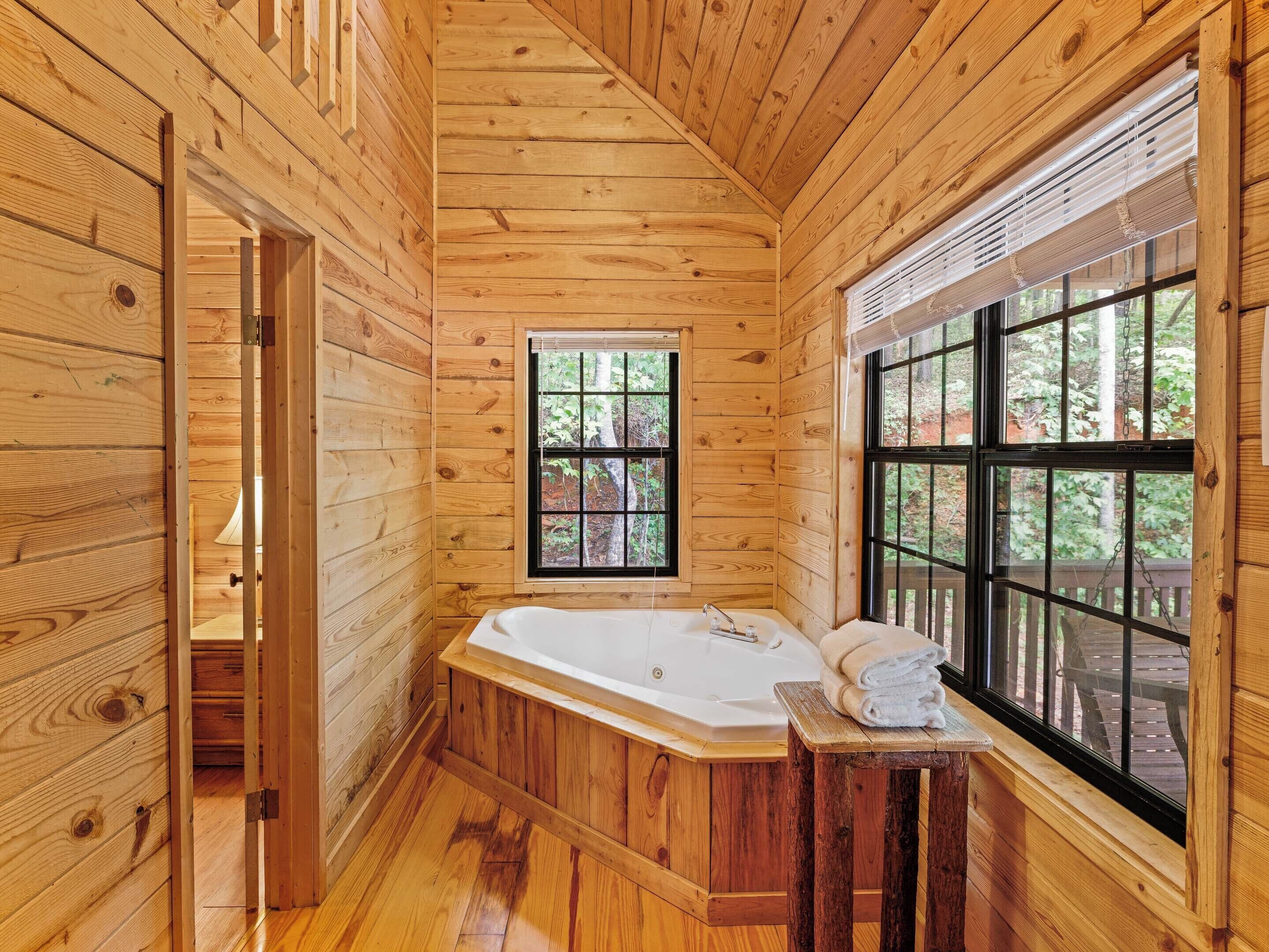 A wooden cabin bathroom with a large window, a bathtub, and a wooden stool with towels.