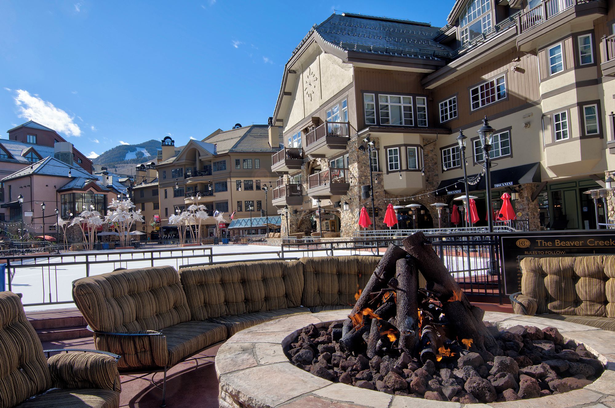 A public square with a stone fire feature, seating areas, and a building with balconies and outdoor seating.