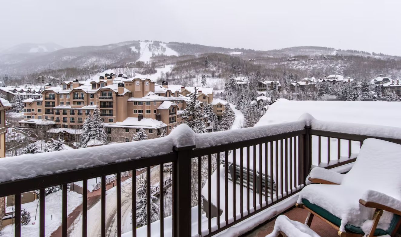 Un balcon enneigé offrant une vue sur un bâtiment d’un complexe hôtelier et sur le paysage enneigé environnant.