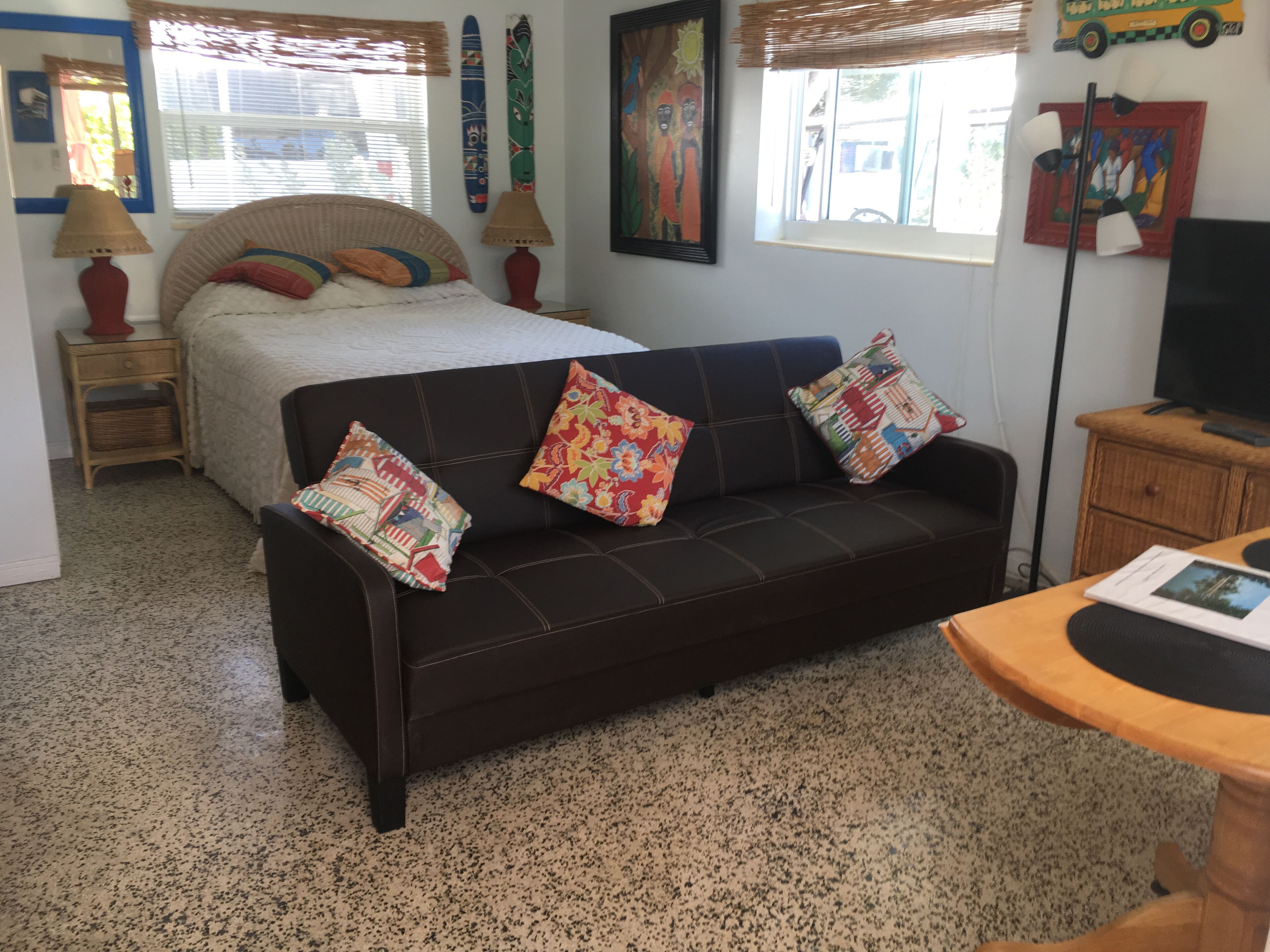 A bedroom with a bed, a dark brown sofa with colorful pillows, a wooden nightstand, a red lamp, and a television on a wooden cabinet.