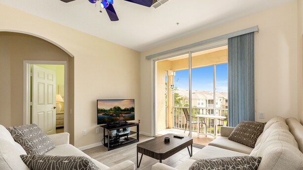 A living room with a flat-screen TV, two sofas, a coffee table, and a sliding glass door leading to a balcony.
