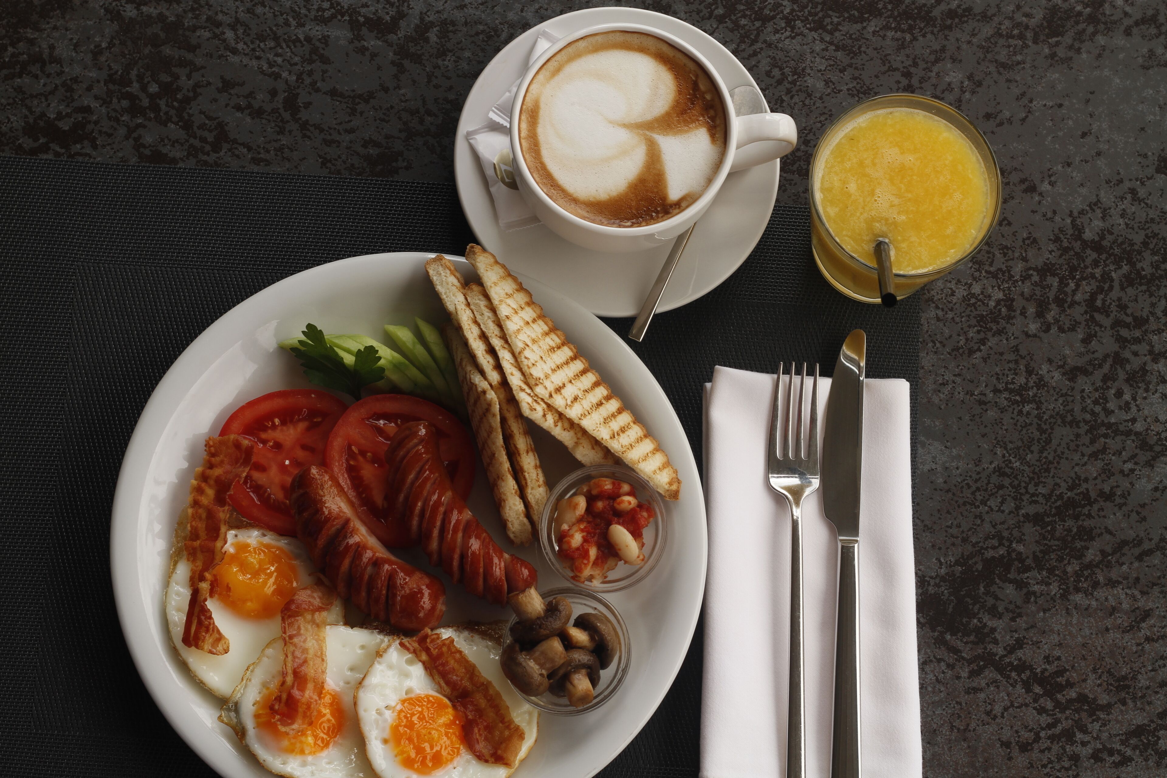 A breakfast plate with eggs, bacon, tomatoes, mushrooms, toast, and a cup of coffee with latte art.