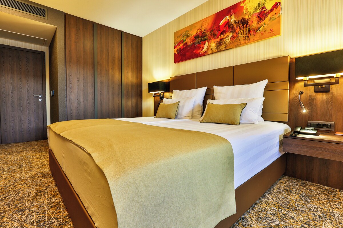 A hotel room with a large bed, a headboard with integrated lighting, a bedside table with a phone, and a painting on the wall.