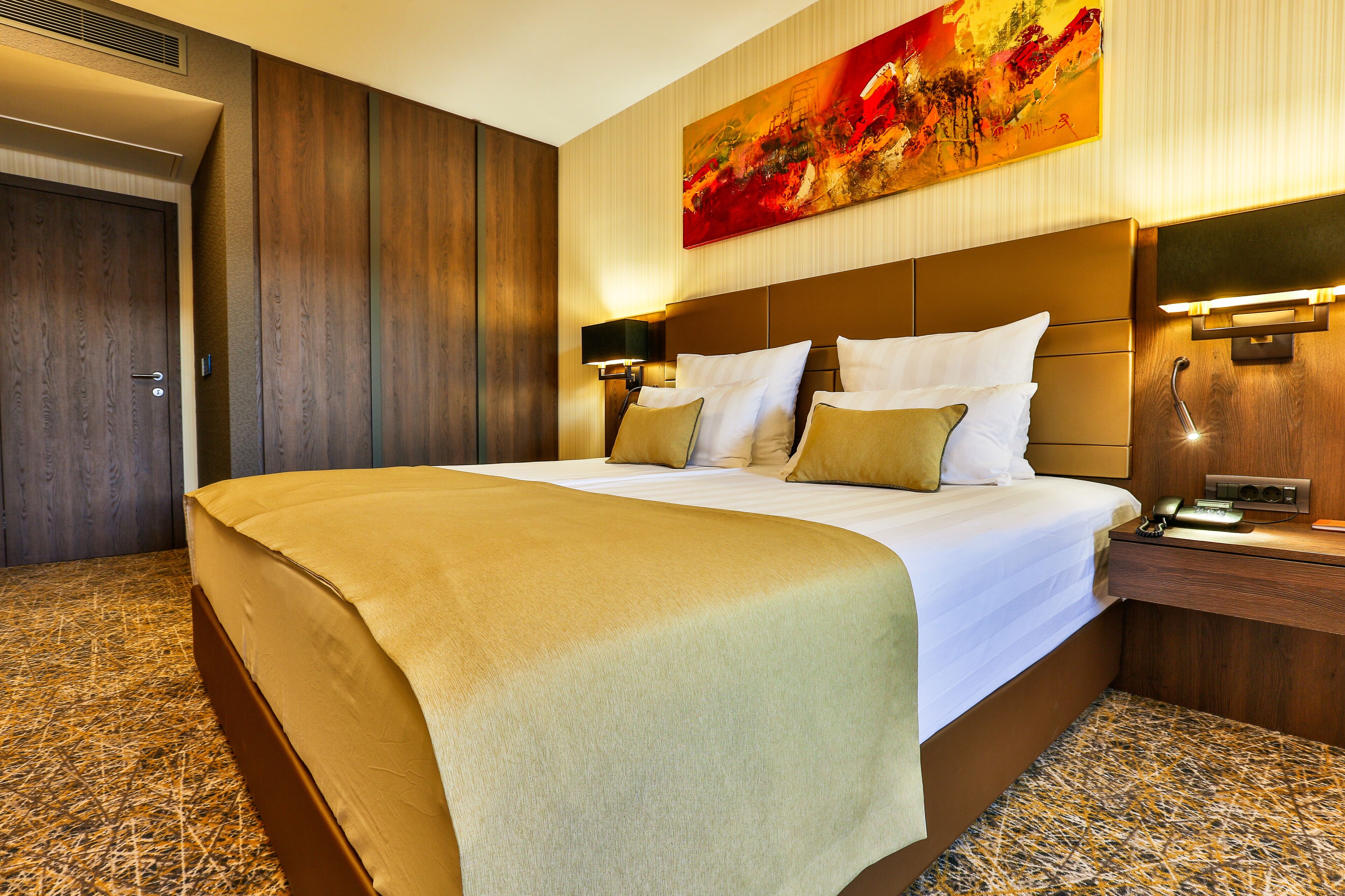 A hotel room with a large bed, a headboard with integrated lighting, a bedside table with a phone, and a painting on the wall.