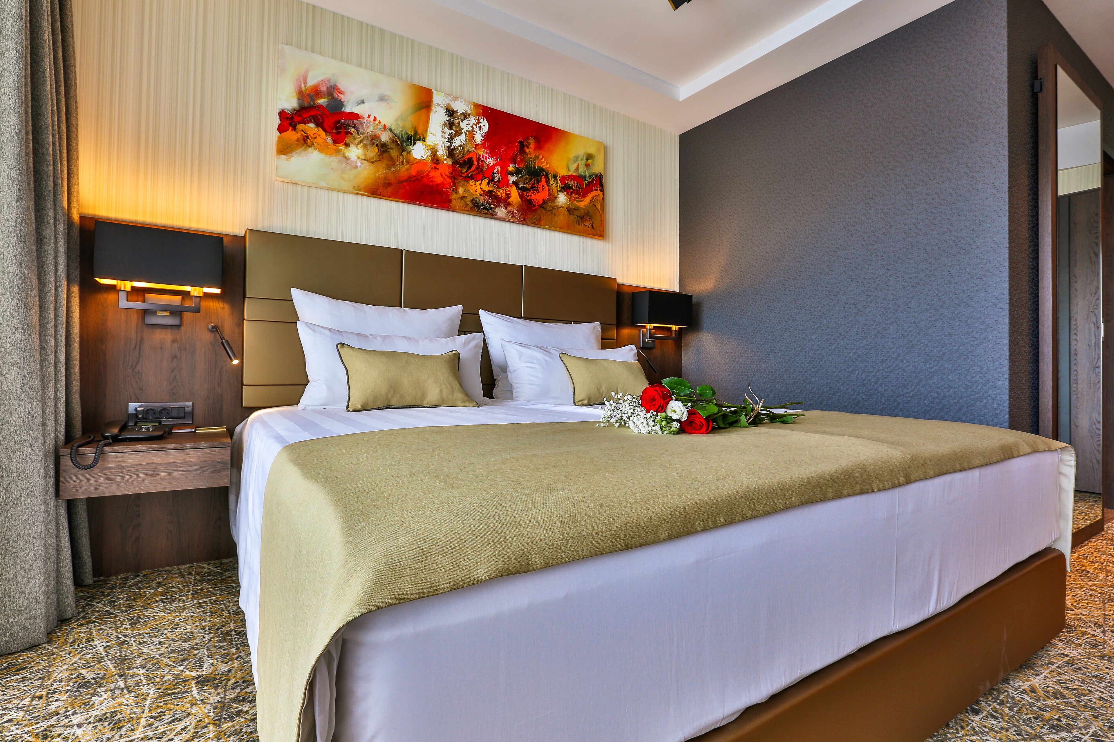 A hotel room with a large bed, a nightstand with a lamp, a painting on the wall, and a floral arrangement on the bed.