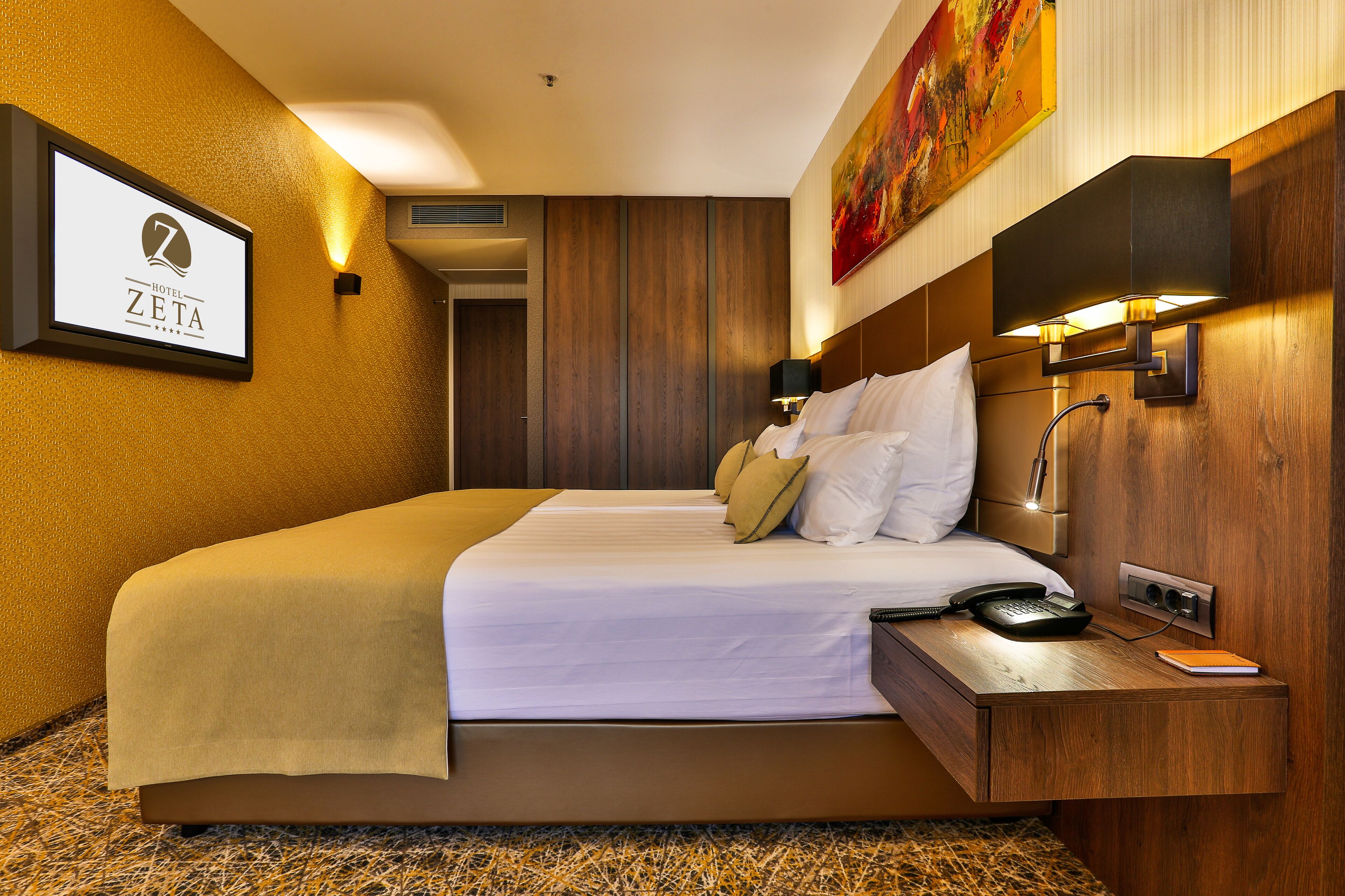 Habitación de hotel con una cama grande, una mesita de noche con lámpara, un cuadro en la pared y un cartel con el nombre del hotel.