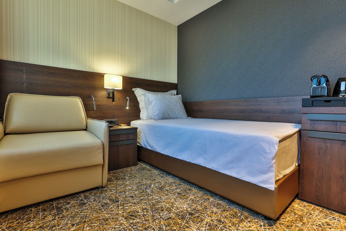 Una habitación de hotel con una cama, una silla, una mesita de noche y una lámpara.