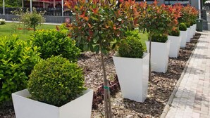 Um edifício moderno com vários andares e varandas, rodeado por um jardim ajardinado com arbustos e árvores diversos.