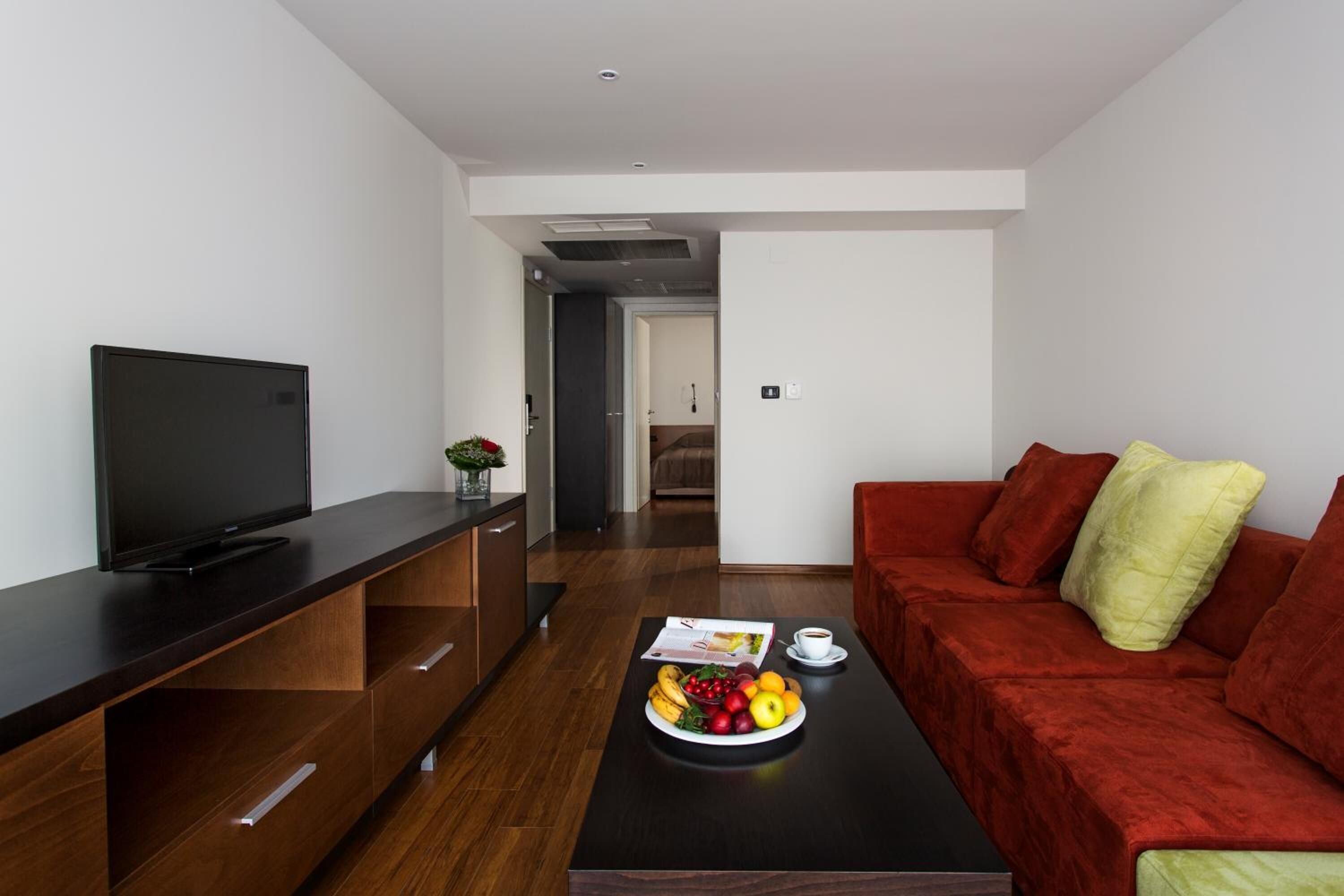 Un salon moderne doté d’un téléviseur à écran plat, d’un meuble de rangement en bois, d’un canapé rouge et d’une table basse sur laquelle sont disposés des fruits.