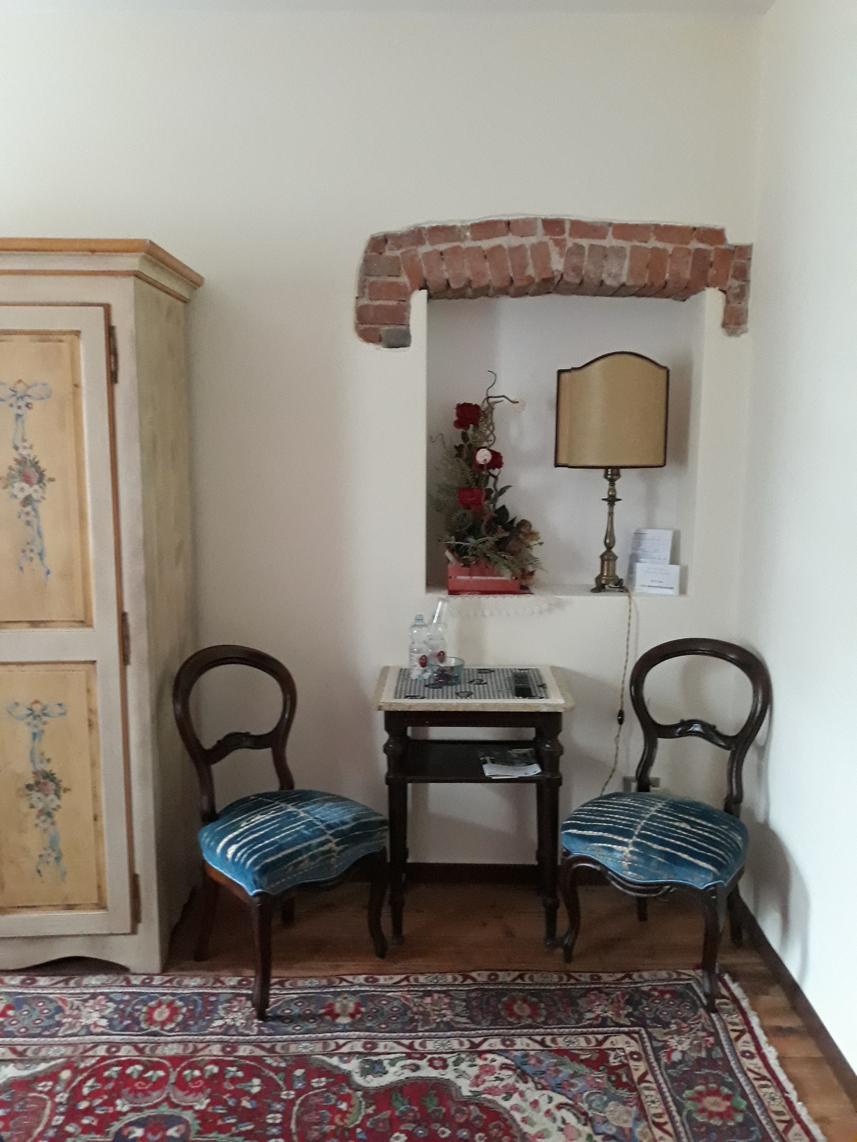 A room with a wooden cabinet, two chairs, a small table with a lamp, a decorative plant, and a patterned rug.