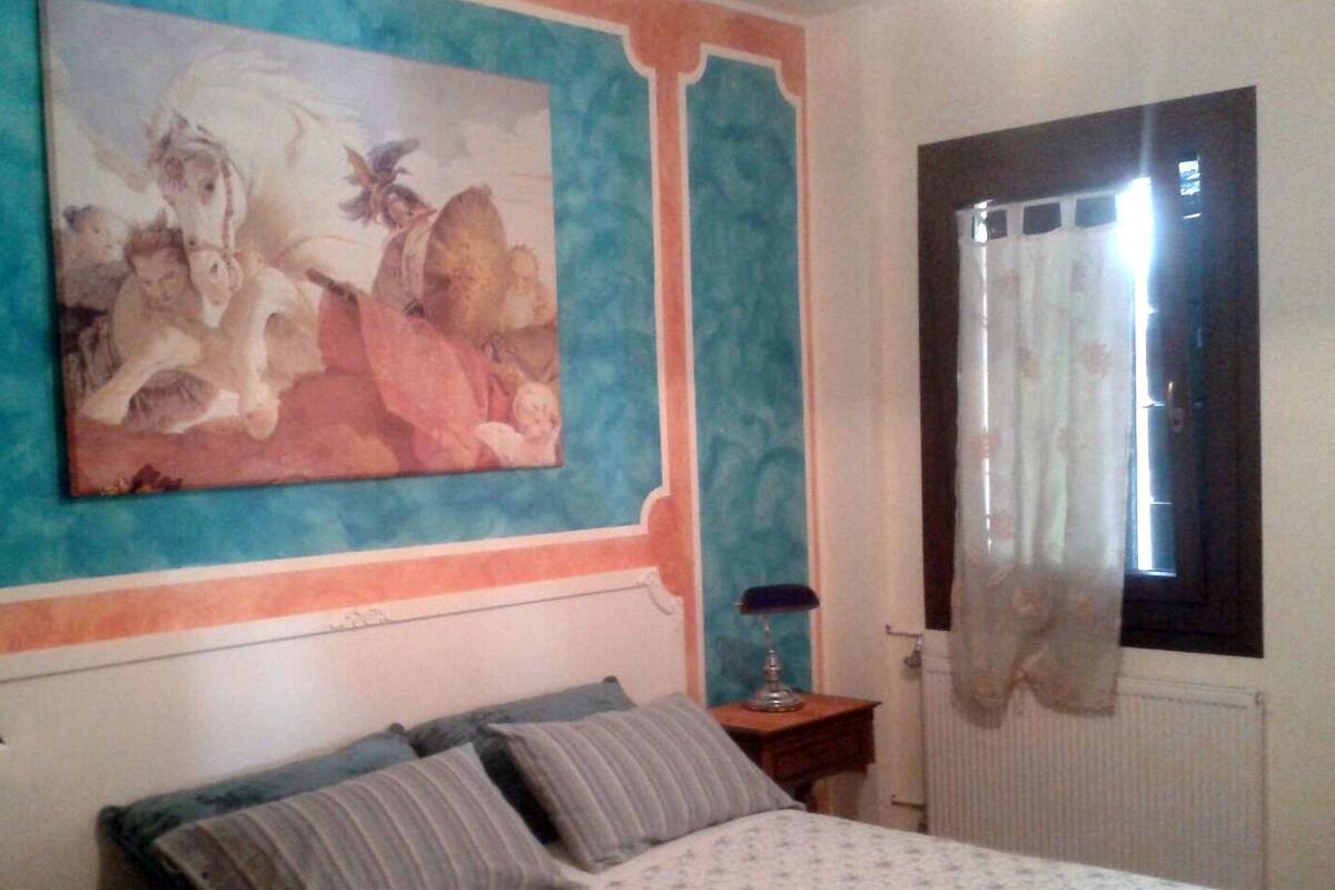 Un dormitorio con cama, almohadas, mesita de noche, lámpara, ventana con cortinas y un cuadro grande en la pared.