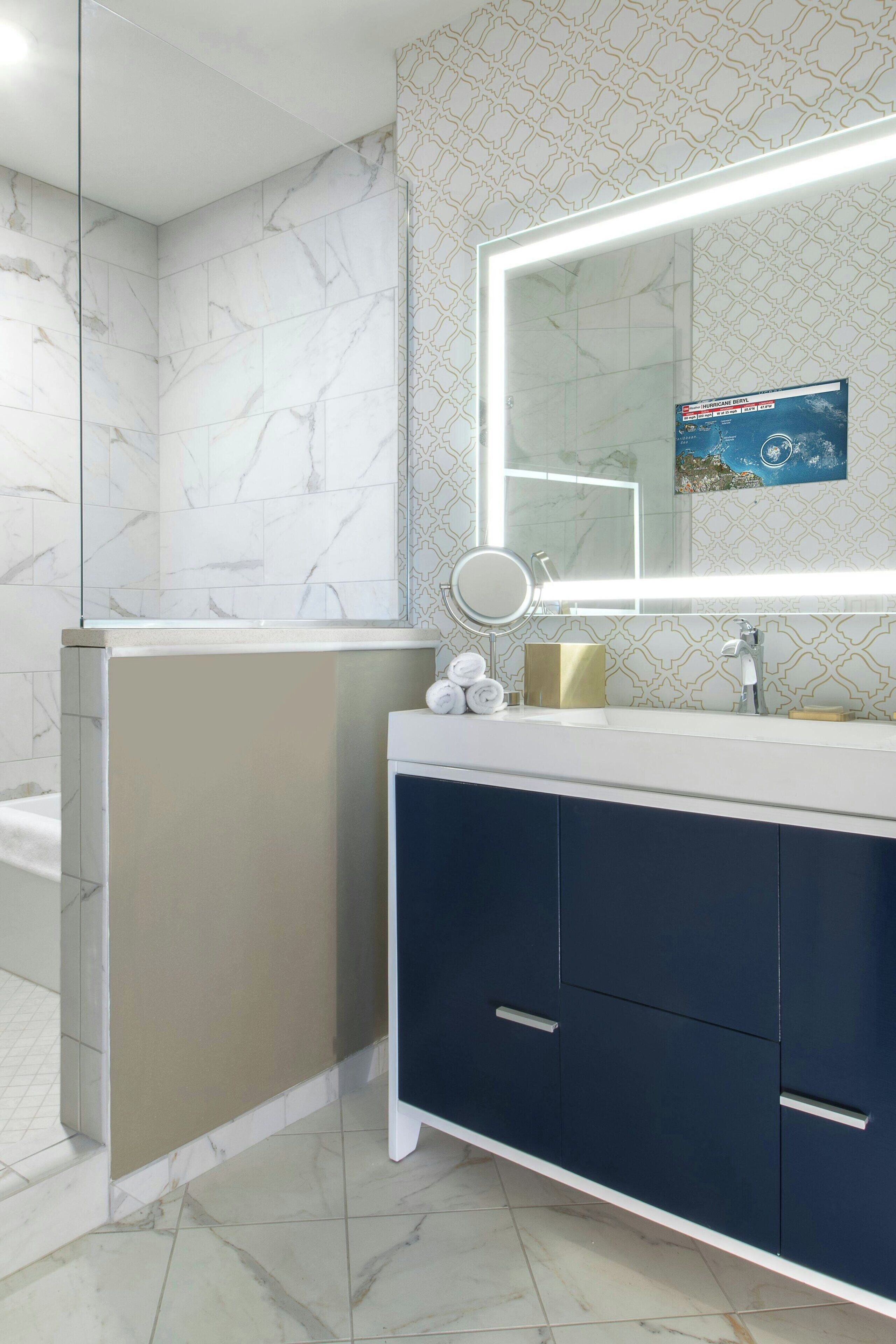 Un baño con pared de azulejos de mármol, un espejo grande con luz, un lavamanos y un mueble de color azul oscuro.