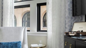 Habitación de hotel con sofá, una mesa redonda con bandeja, cómoda negra y un ventanal con vista a un edificio.