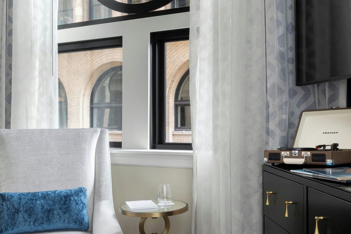 A hotel room with a sofa, a round table with a tray, a black dresser, and a large window with a view of a building.