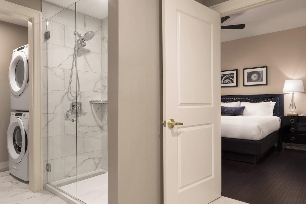 A hotel room with a glass-enclosed shower, a bed with dark wood headboard, and a bedside table with a lamp.