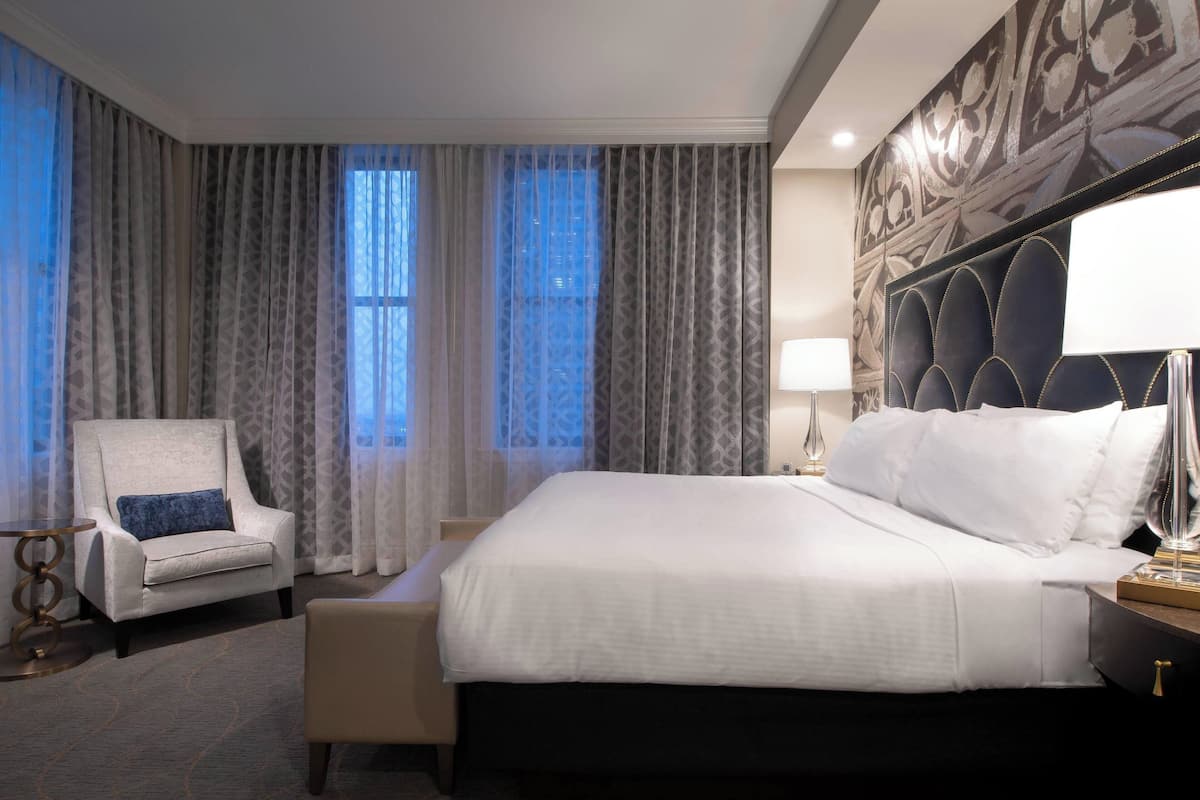 A hotel room with a large bed, a chair, a small table, and a decorative headboard.
