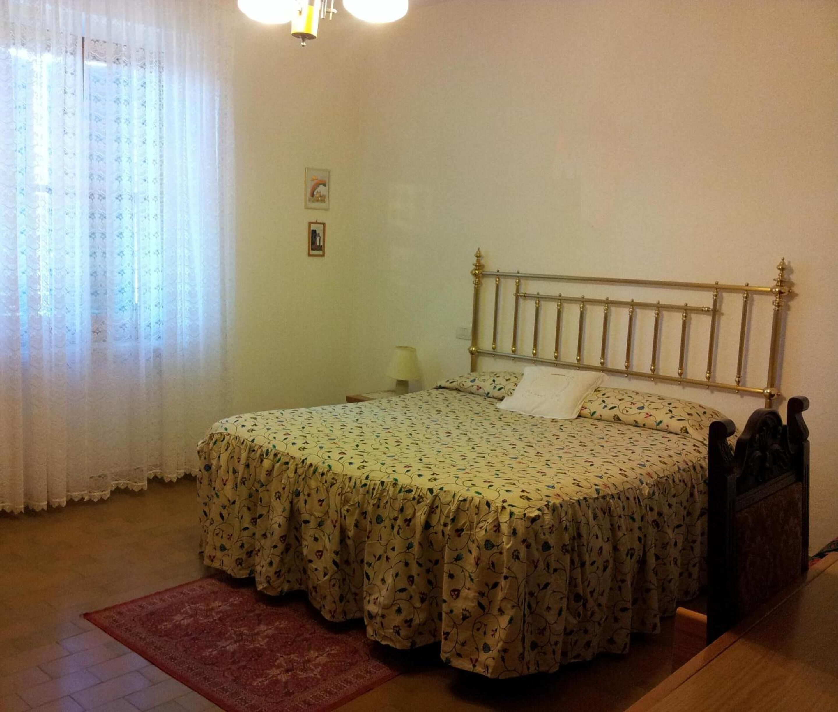 A bedroom with a bed, a nightstand, a lamp, a window with curtains, and a wall with a picture.