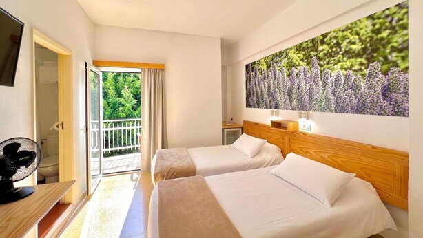 Habitación de hotel con dos camas, un balcón, un ventanal grande con vistas a la naturaleza y un ventilador a la izquierda.