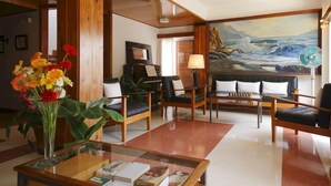 Un hall con mobiliario de madera, un cuadro grande de un paisaje marino y un ventilador.