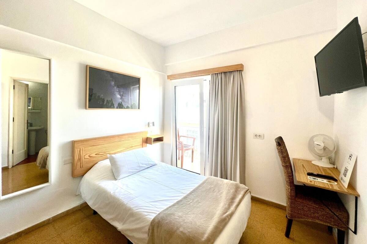 Habitación de hotel con cama, una silla, un escritorio con lámpara, televisor y ventana con cortinas.