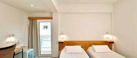 A hotel room with two single beds, a small desk with a chair, a fan, and a view of the outside through a door.