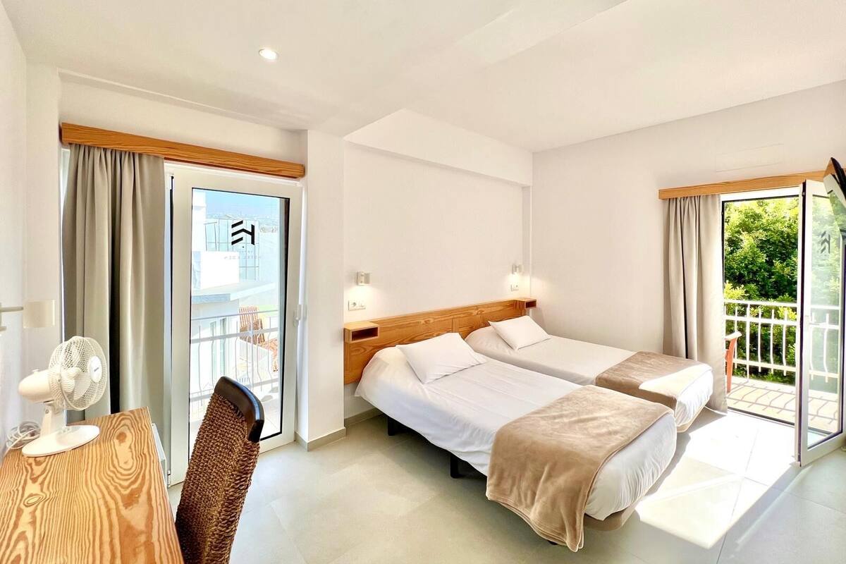 Habitación de hotel con dos camas, un escritorio con un ventilador y vistas a zonas verdes a través de las ventanas.