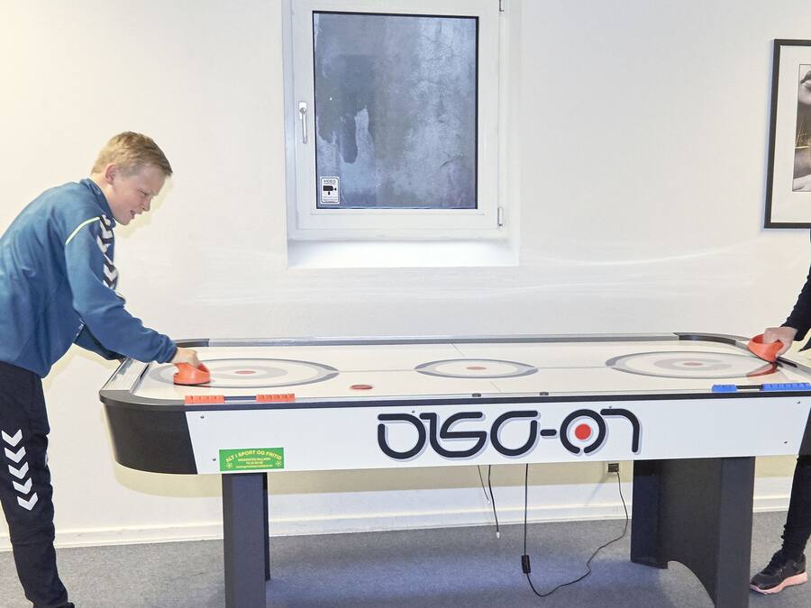 Two people playing table tennis in a room with a table tennis table, a window, and a framed picture on the wall.
