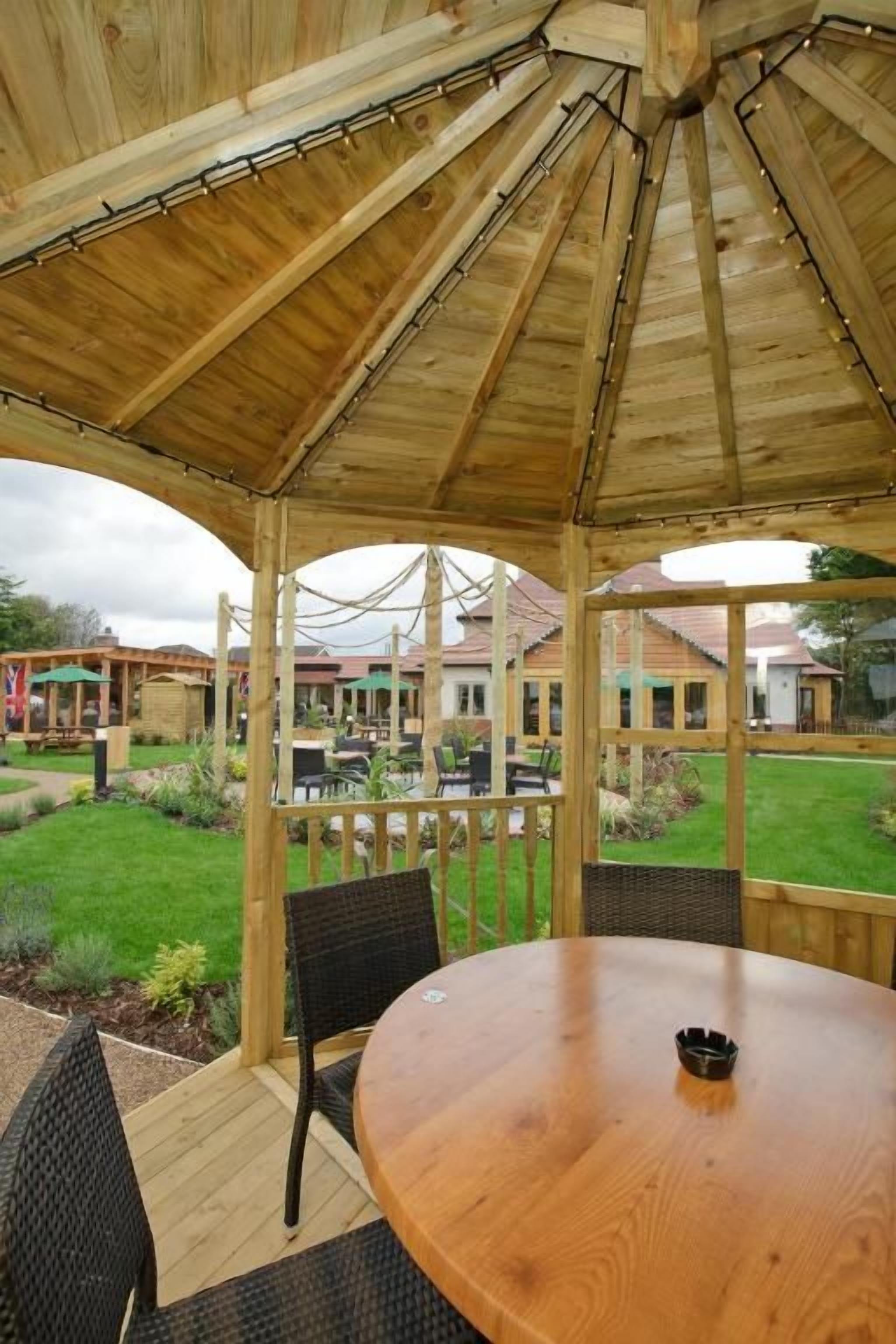 Um gazebo de madeira com uma mesa redonda e cadeiras, cercado por um deck de madeira e uma área gramada.
