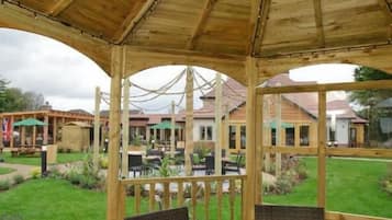 Um gazebo de madeira com uma mesa redonda e cadeiras, cercado por um deck de madeira e uma área gramada.