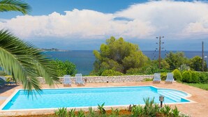 A rectangular swimming pool with a stone border, surrounded by lounge chairs and greenery, overlooking a body of water and a distant coastline. - Akritas (Pylos-Nestoras)