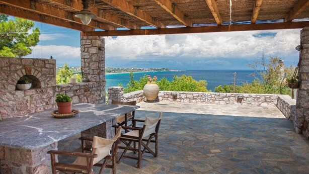 An outdoor patio with stone walls, a stone table, and chairs, overlooking a scenic coastal view. - Akritas (Pylos-Nestoras)