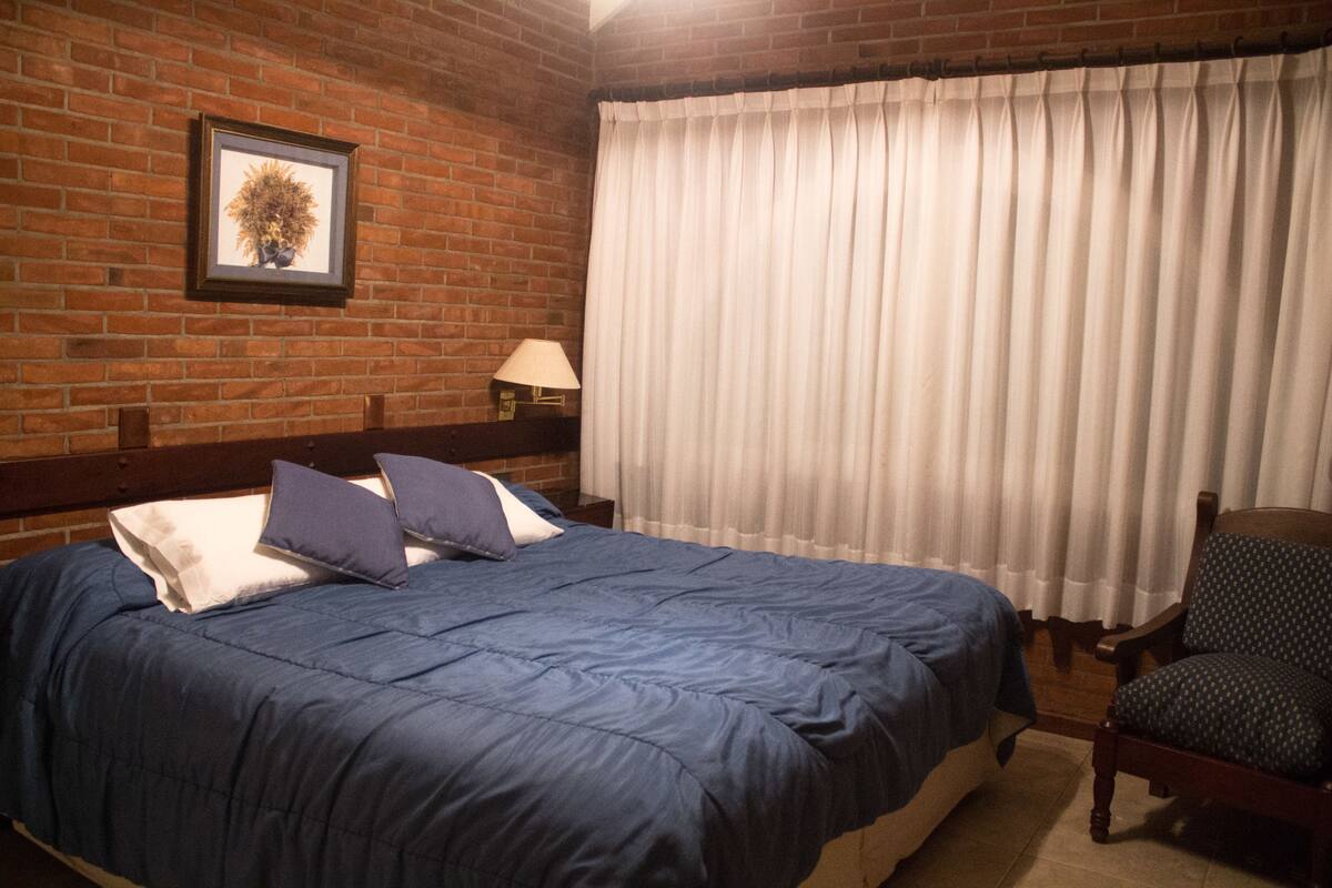 A bedroom with a brick wall, a framed picture, a bed with blue bedding, a chair, and a window with white curtains.