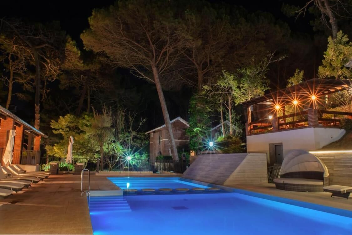Zona de piscina de nit amb gandules i una zona d'estar coberta, envoltada d'arbres i un edifici amb finestres il·luminades.