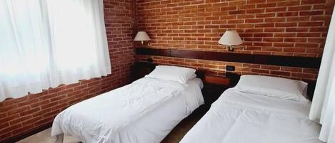 Una habitación con dos camas, una pared de ladrillo y una ventana con cortinas blancas.
