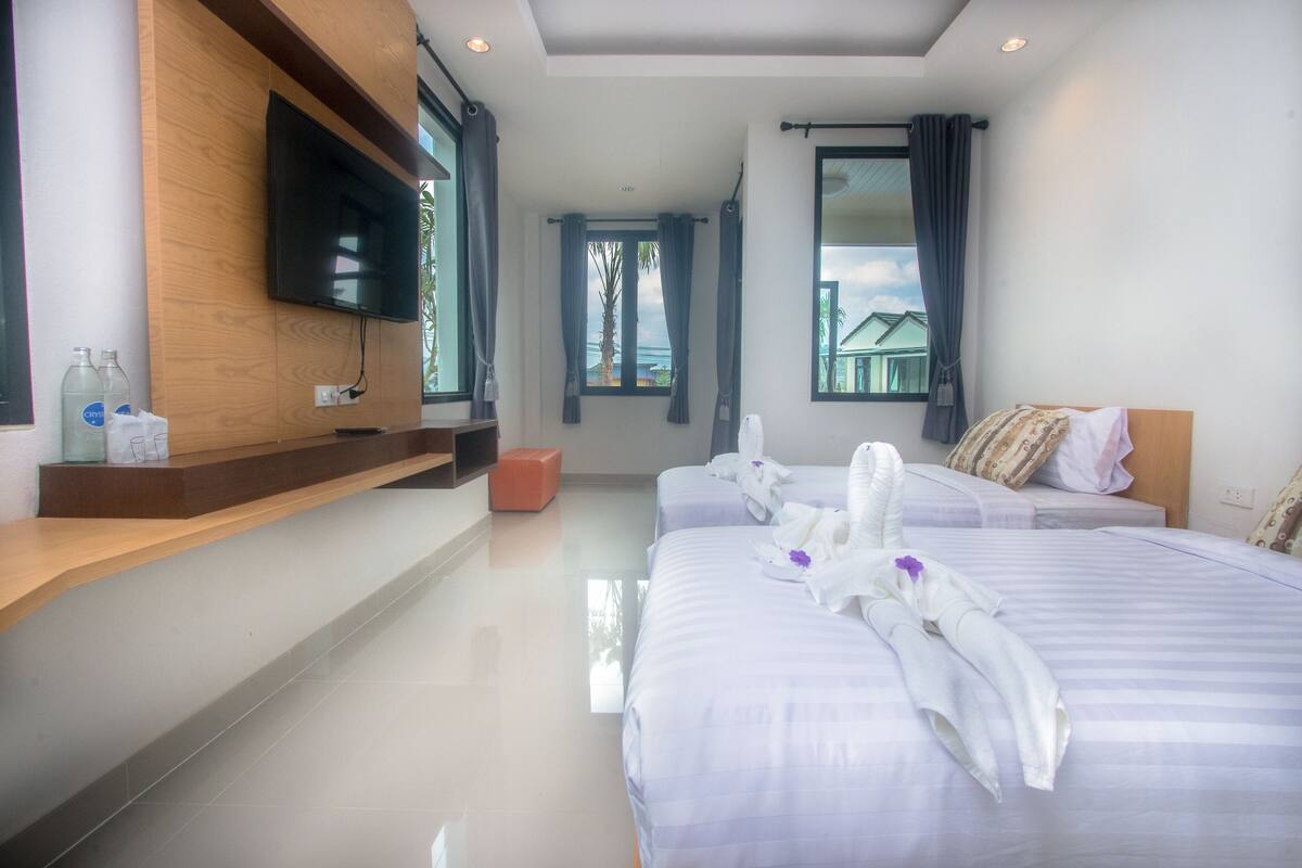 A hotel room with two beds, a TV mounted on the wall, a wooden shelf with bottles, and a window with curtains.
