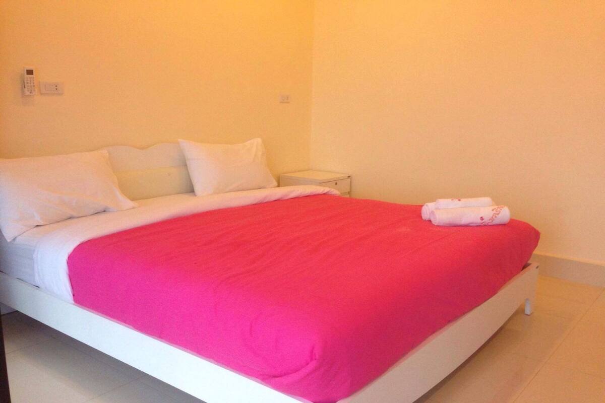 Una cama con ropa blanca y una manta rosa brillante, una mesita de noche blanca y una pared blanca con un interruptor de luz.