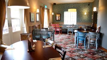 A dining area with a round table, chairs, and a patterned carpet. There are windows with curtains, wall-mounted lamps, and a small table with a chair.