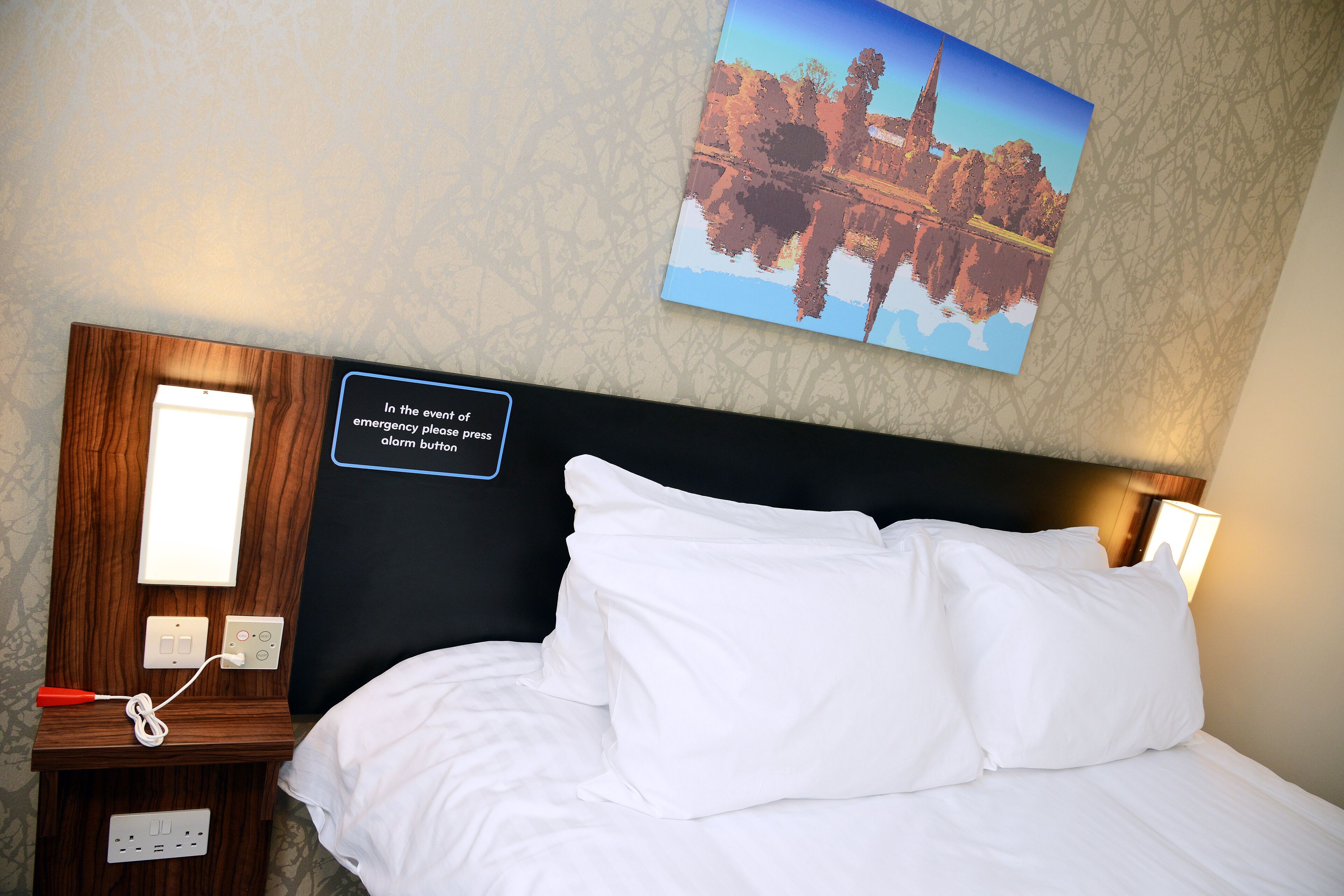 A neatly made bed with white linens, a wooden nightstand with a lamp, and a wall-mounted emergency instruction sign.