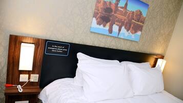 A neatly made bed with white linens, a wooden nightstand with a lamp, and a wall-mounted emergency instruction sign.