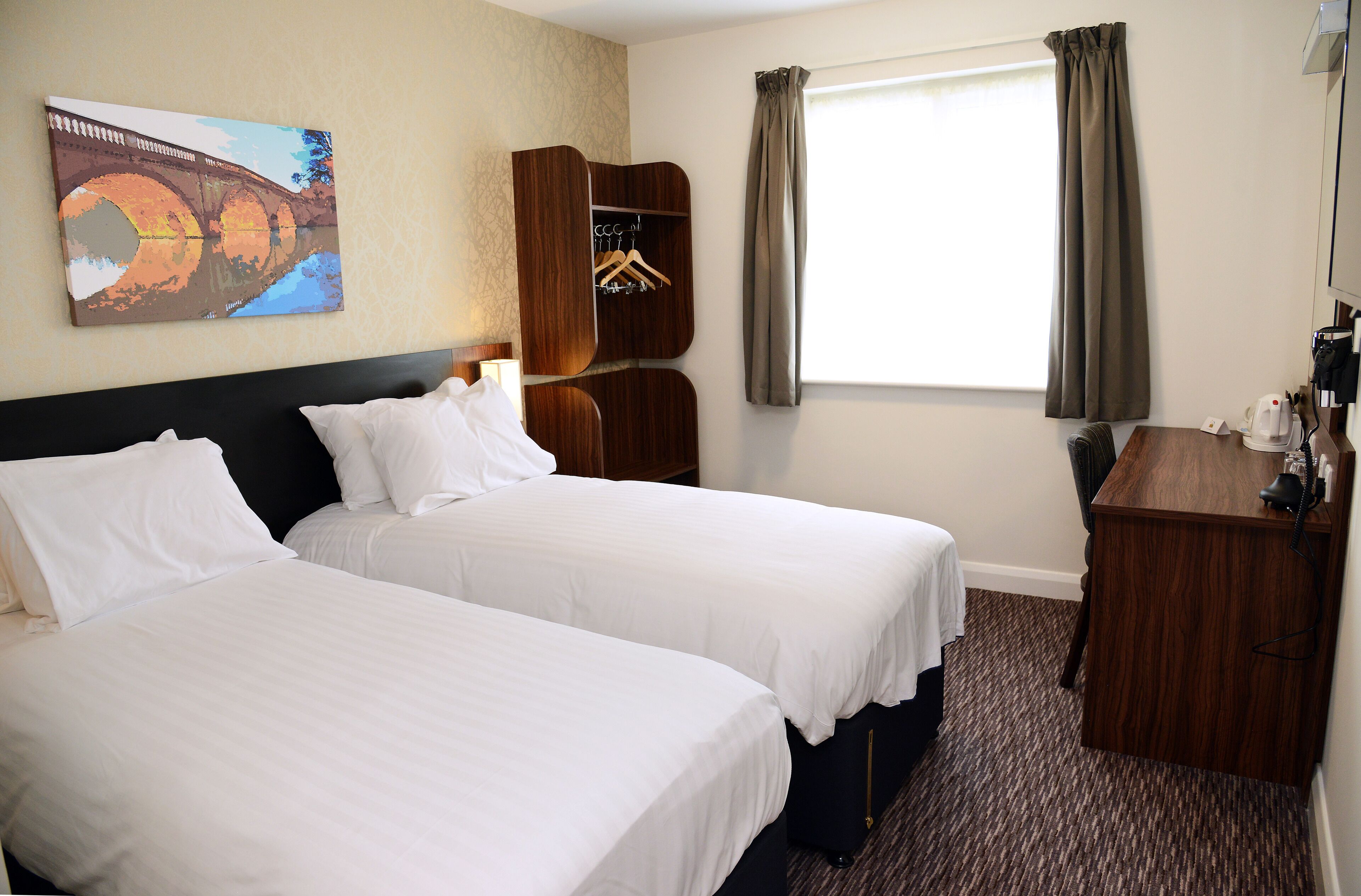 A hotel room with two beds, a wooden wardrobe, a coat rack, and a painting on the wall.