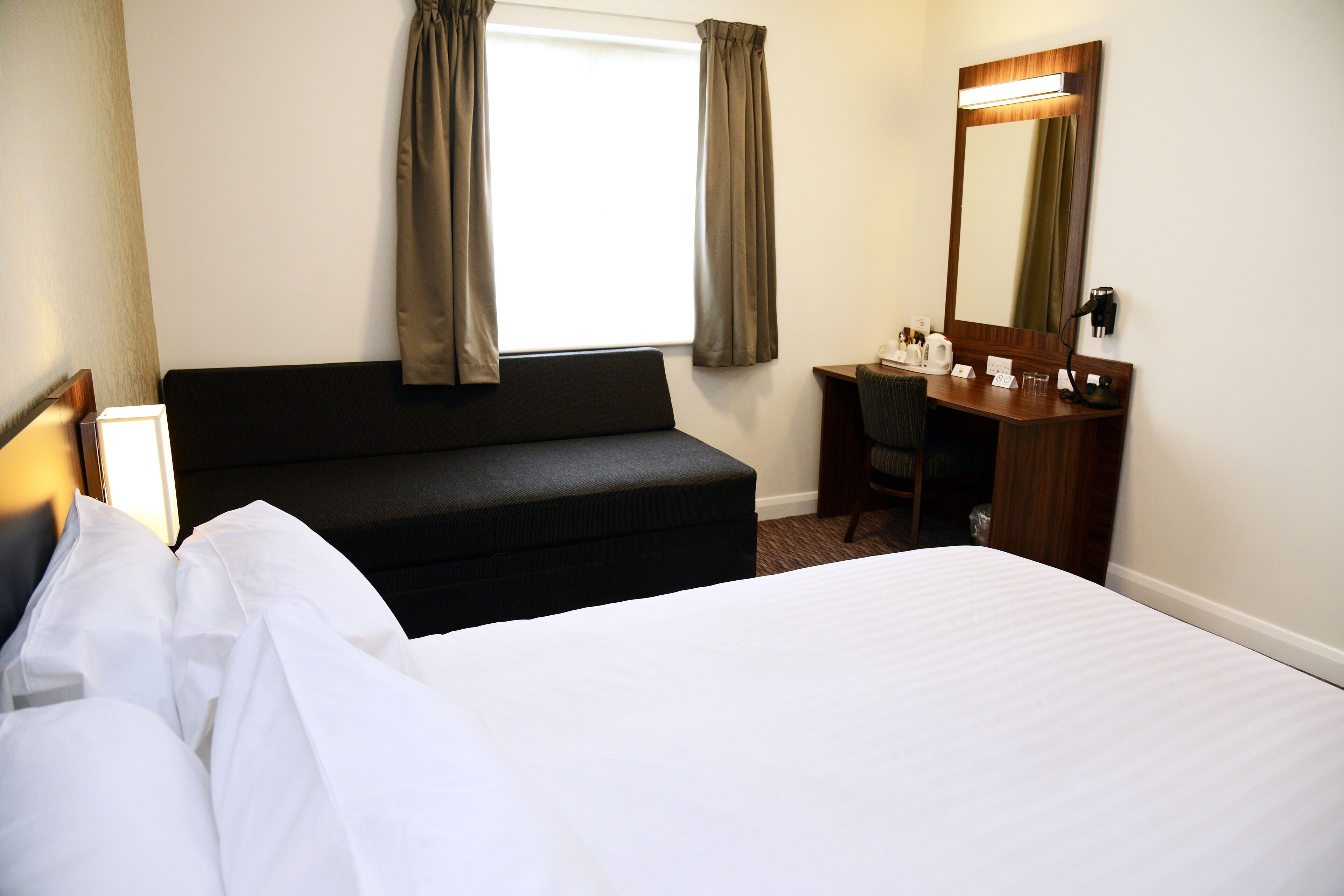 A hotel room with a bed, a desk, a chair, a mirror, and a window with curtains.