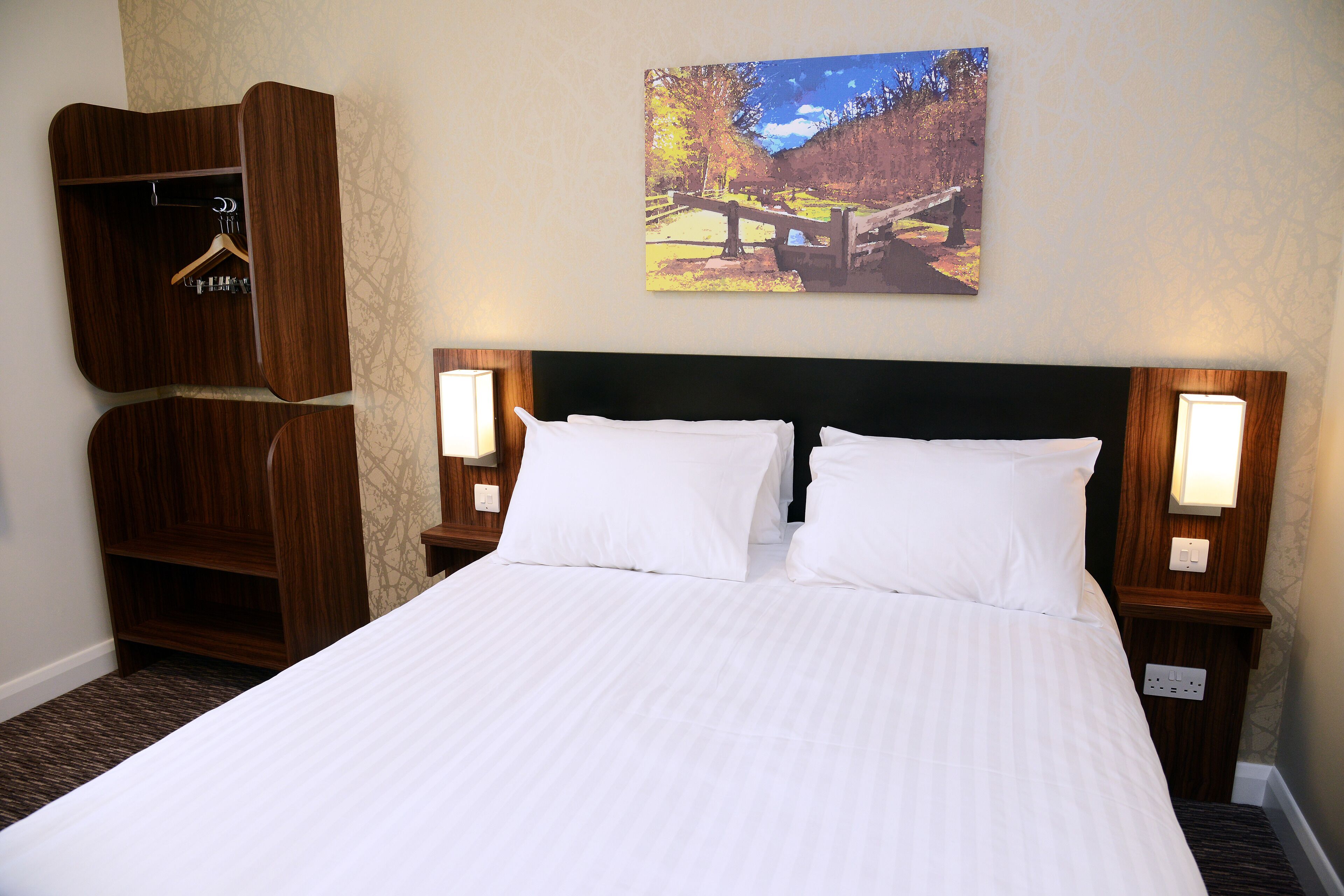 A hotel room with a large bed, wooden wardrobe, and a painting of a scenic landscape on the wall.