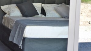 Una cama con cabecera gris y ropa de cama blanca, ubicada en una habitación con pared amarilla y ventana con vistas a vegetación.